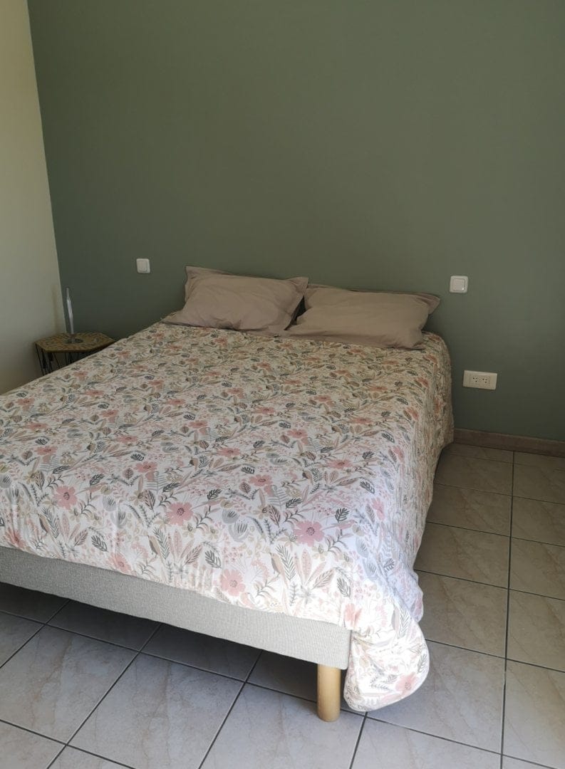 Chambre avec un lit double, couvert d'une couette à motifs floraux, et deux oreillers beiges. Le mur est peint en vert avec des prises de courant visibles.