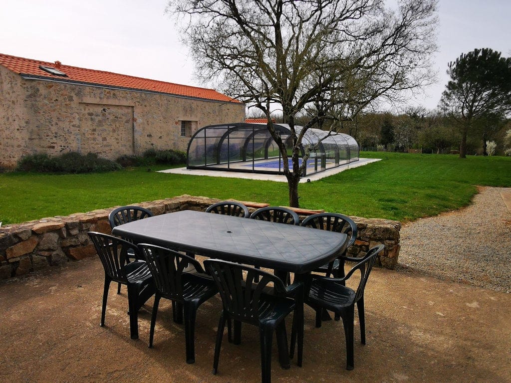 Espace extérieur du Gîte Le Graizeau avec une table et des chaises en plein air, offrant une vue sur la piscine couverte et le jardin verdoyant.