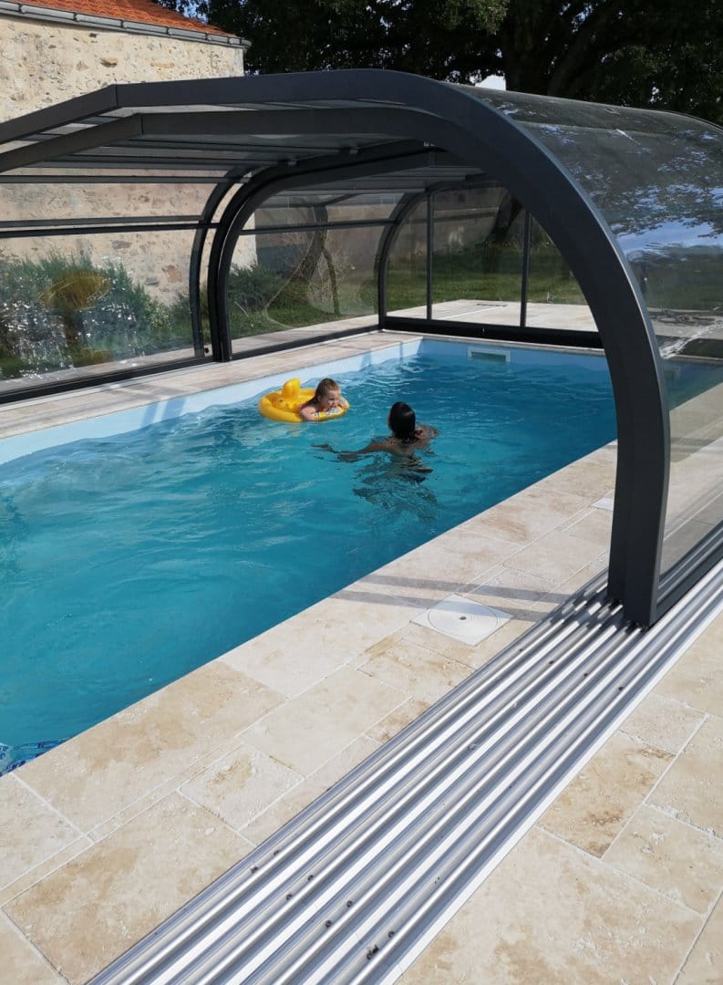 Piscine familiale couverte par un abri en verre et métal, avec deux personnes profitant de l'eau, entourée de dalles en pierre type travertin